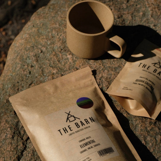 A gift box setup with a bag of coffee beans labeled 'The Barn', a beige Hasami mug, and a small bag of Cascara, against a background of natural stone.