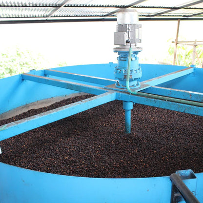 Blue cylindrical machine with a central shaft processing dark material, likely coffee beans, outdoors.