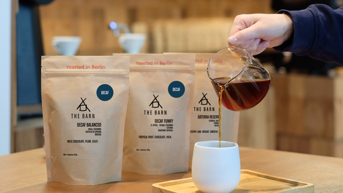 Person pouring coffee from a glass server into a white cup with coffee bags labeled 'The Barn' in the foreground.