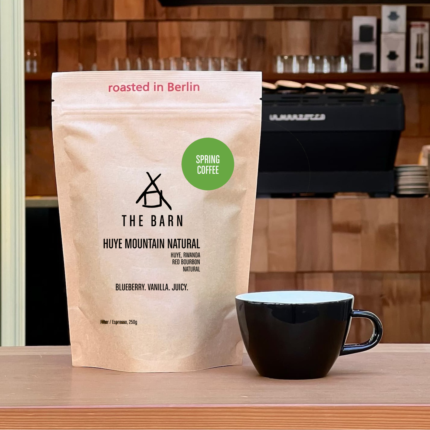 Spring Coffee bag labeled 'Huye Mountain Natural' and a black coffee cup on a wooden surface with a kitchen background.