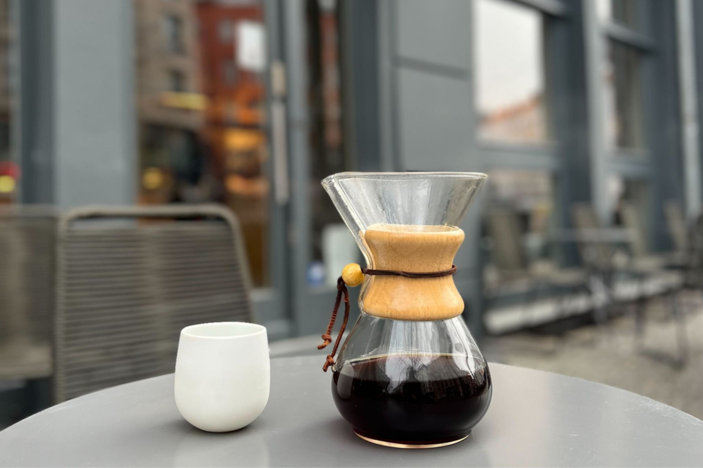 A Chemex glass coffee maker filled with freshly roasted coffee, with a ceramic mug on a table