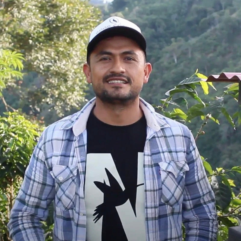 Coffee Farmer wearing a plaid shirt over a black t-shirt with a graphic design, standing outdoors with greenery in the background.
