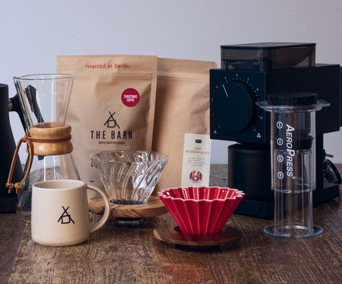 A collection of curated coffee gifts on a wooden table consists of a bag of coffee, a opus grinder, a clear aeropress a sandstone mug and origami drippers.
